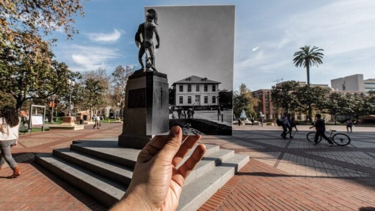 A hand holding out an old photograph of campus with the Tommy Trojan statue in the exact area on campus in the modern day.
